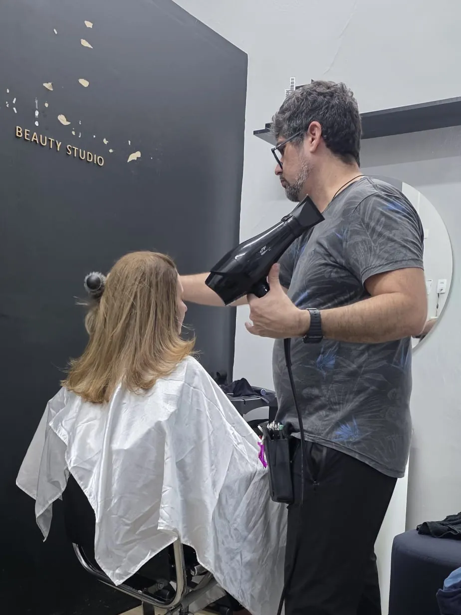 Pablo seca com secador o cabelo loiro de uma cliente de capa branca, diante da parede preta do estúdio