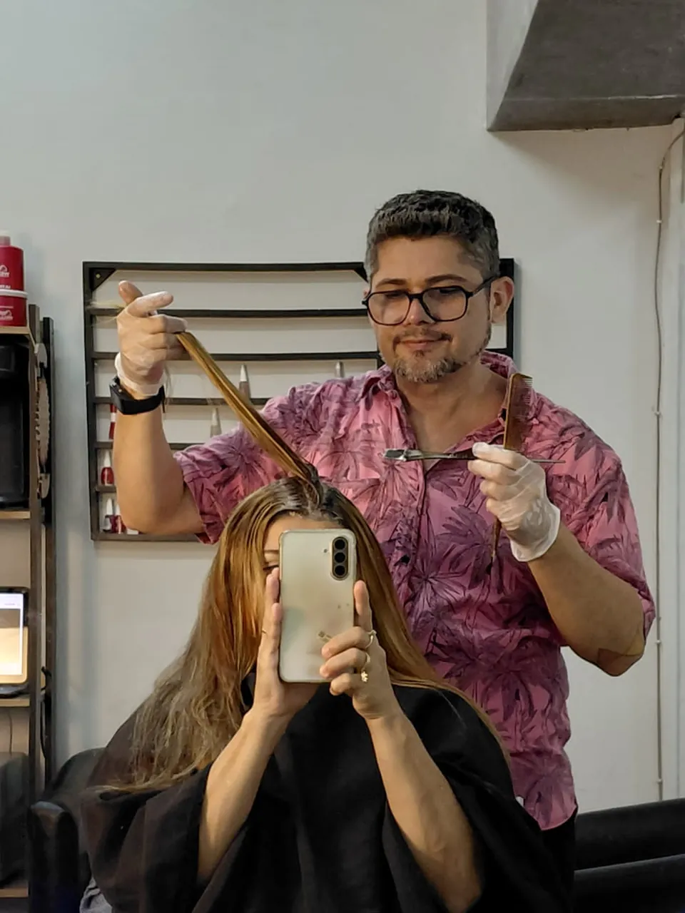 Pablo, de camisa estampada e luvas, ergue uma mecha para avaliar o comprimento enquanto a cliente fotografa no espelho